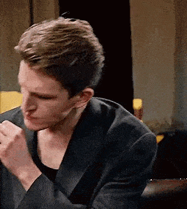 a man in a suit is sitting on a couch with his hands folded in front of him .