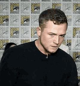 a man in a black shirt is standing in front of a wall with comic con logos on it .