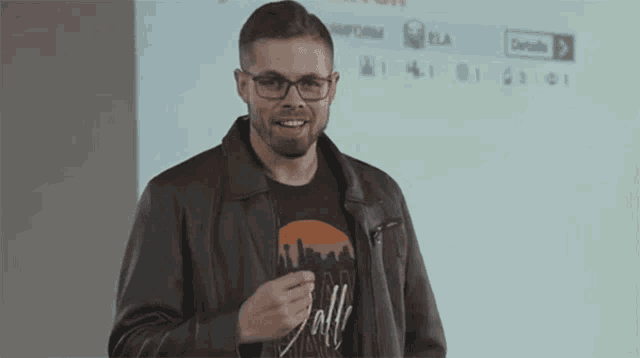 a man wearing glasses and a leather jacket is giving a thumbs up while standing in front of a projection screen .