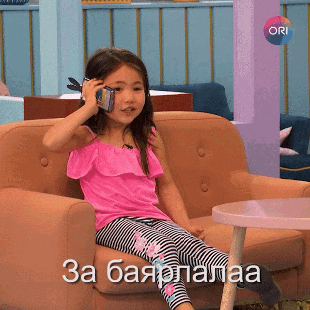a little girl sitting on a couch talking on a cell phone with ori in the corner