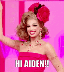 a drag queen wearing a red dress and a flower in her hair says hi aiden