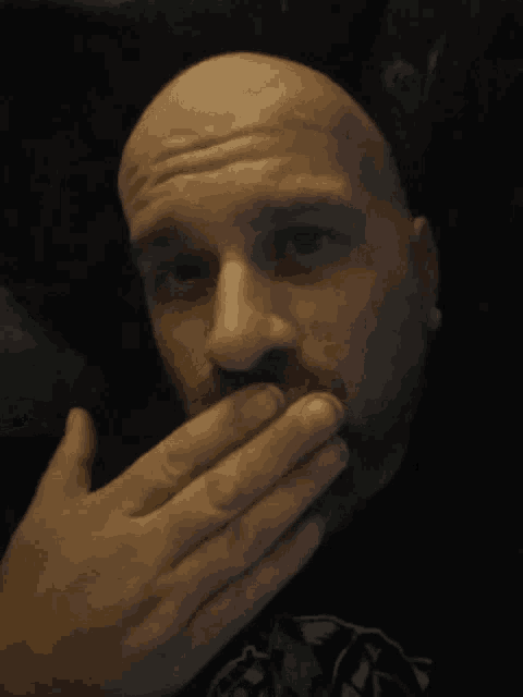 a bald man covering his mouth with his hand in a dark room