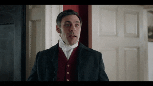 a man in a black suit and red vest is standing in a doorway