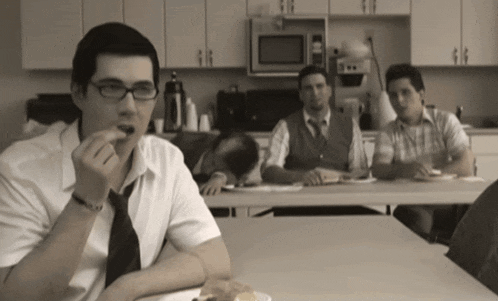 a man wearing glasses sits at a table eating a snack