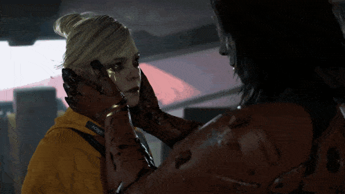 a woman wearing a yellow jacket with the word joker on it is touching another woman 's face
