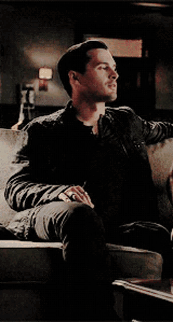 a man in a black jacket is sitting on a couch with his legs crossed