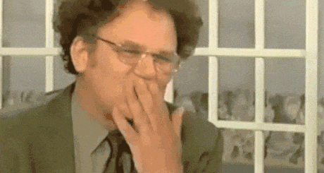 a man in a suit and tie is covering his mouth with his hand while sitting in front of a window .