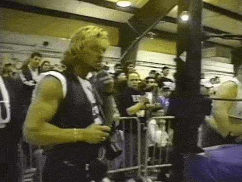 a man in a black vest with the letter e on it stands in front of a crowd holding a camera .