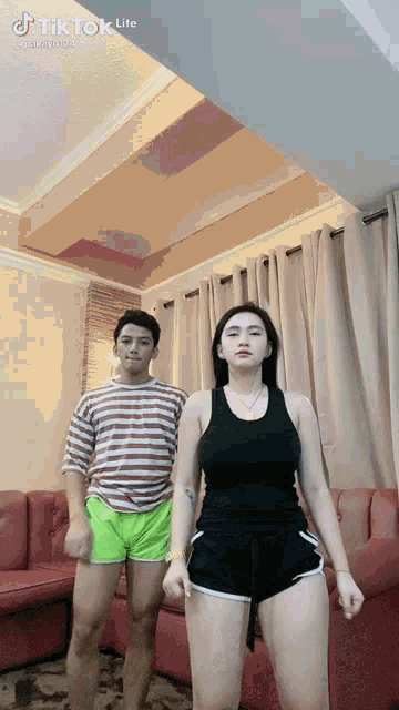 a man and a woman are standing next to each other in a living room . the woman is wearing shorts .