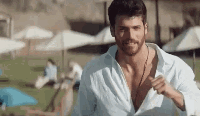 a man with a beard wearing a white shirt and a necklace is standing in front of a beach .