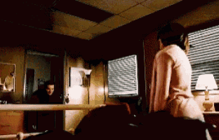 a woman in a pink sweater is standing in a dark room with blinds on the windows .