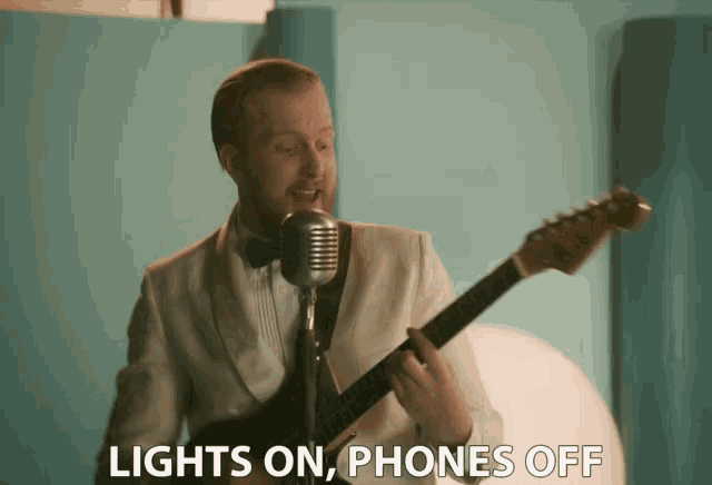 a man singing into a microphone while holding a guitar with the words lights on phones off behind him