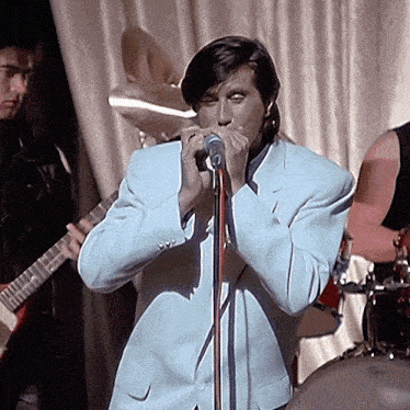 a man singing into a microphone while another man plays a guitar in the background