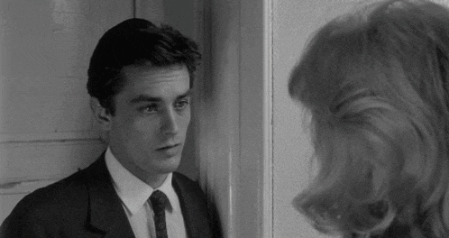 a man in a suit and tie leans against a wall looking at a woman