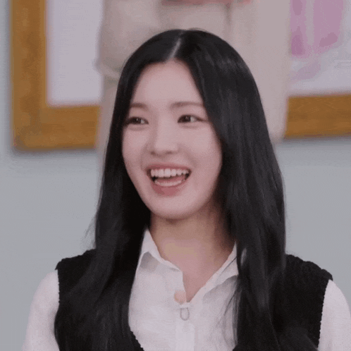 a young woman with long black hair is smiling and wearing a white shirt and a black vest .