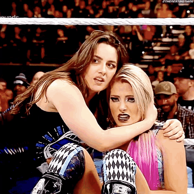 two female wrestlers are hugging each other in a wrestling ring with the next thing written on the bottom right