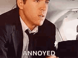 a man in a suit and tie is sitting in the back seat of an airplane and says `` annoyed '' .