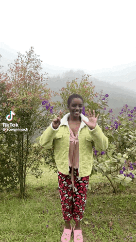 a woman in a green jacket and pink pants is standing in front of purple flowers