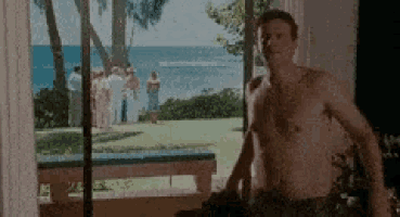 a man without a shirt is standing in front of a window overlooking the ocean .