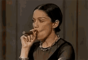 a woman is smoking a cigar while wearing a black dress and a necklace .
