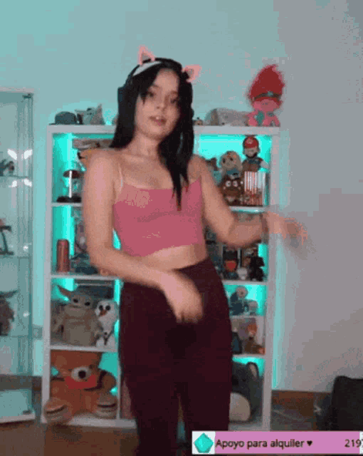 a woman in a pink crop top and purple pants is dancing in front of a shelf with stuffed animals on it