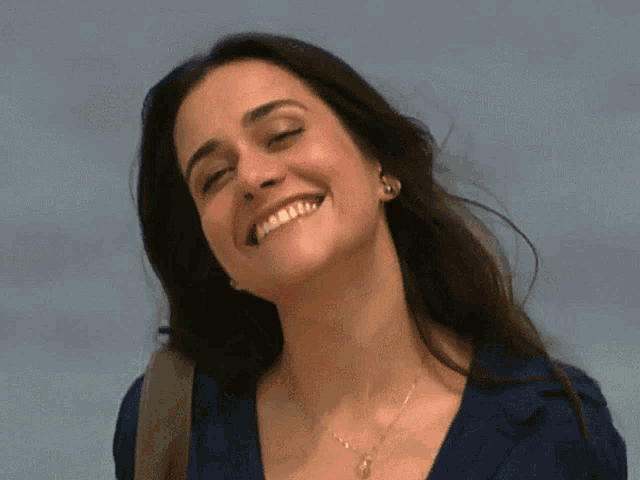a woman wearing a blue shirt and a necklace smiles with her eyes closed