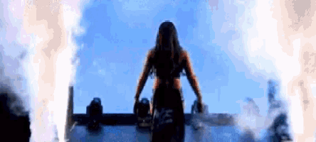 a woman is standing on a stage in front of a blue sky with fire coming out of it .