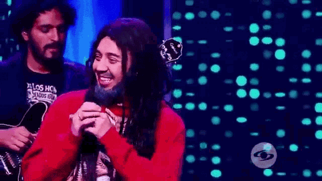 a man with long hair and a beard is singing into a microphone while another man plays a guitar .