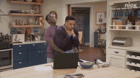 two men are standing in a kitchen looking at a laptop with the nick logo on the shelf