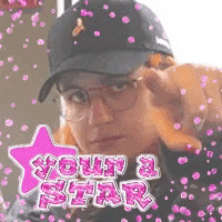 a woman wearing a hat and glasses is pointing at the camera with a pink star in the background .