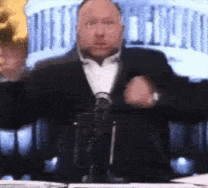 a man in a suit and tie is sitting at a table with a microphone .