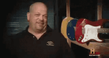 a bald man is holding a red electric guitar in front of a row of other guitars .
