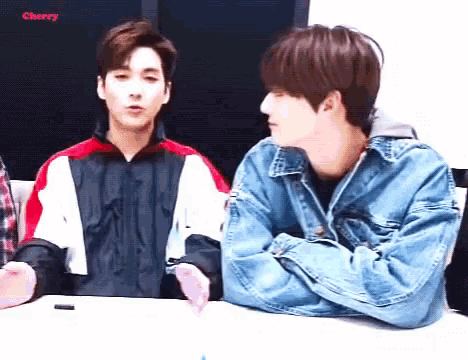 two young men are sitting at a table talking to each other .