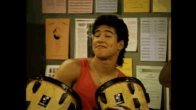 a man in a red tank top is playing drums in front of a wall with posters on it