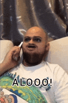 a bald man with a beard is sitting on a couch talking on a cell phone with the words alooo written below him