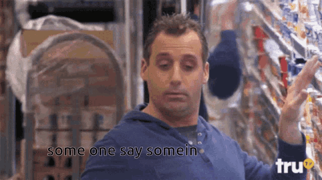 a man in a blue shirt is standing in front of a grocery shelf and says some one say somein