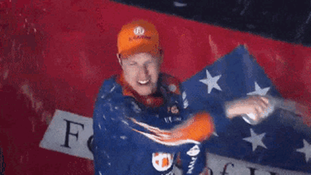 a man in an orange hat stands in front of an american flag with the letter f on it