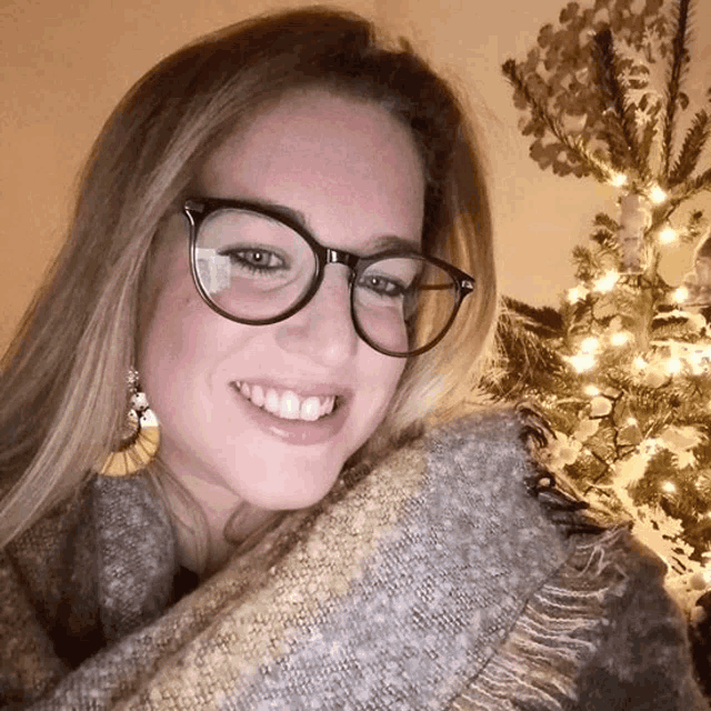 a woman wearing glasses and a scarf stands in front of a christmas tree