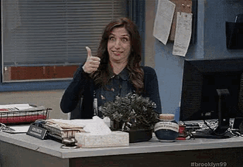a woman is giving a thumbs up while sitting at a desk in front of a computer monitor .