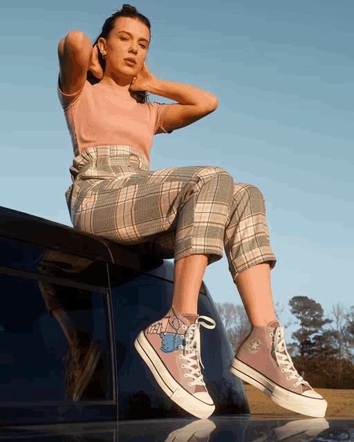 a woman in plaid pants sits on the roof of a car
