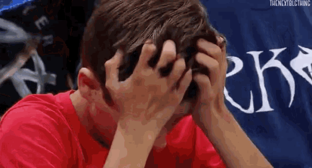 a man in a red shirt is covering his face with his hands in front of a sign .