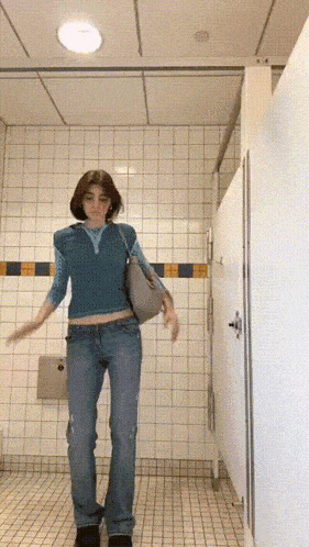 a woman in a blue top and jeans is standing in a public restroom .
