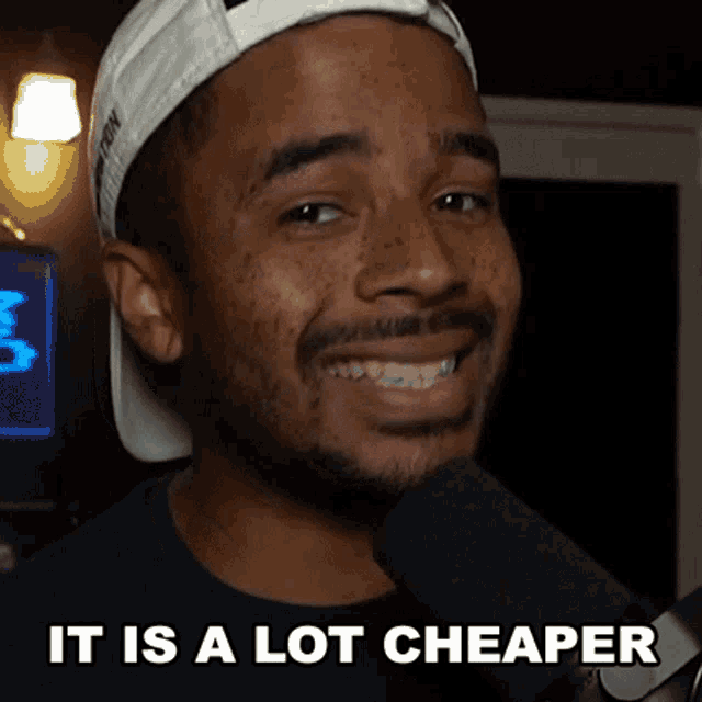 a man wearing a hat is smiling in front of a microphone with the words " it is a lot cheaper " above him
