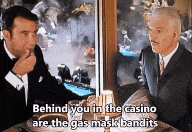 two men are sitting at a table in a casino and behind you in the casino are the gas mask bandits .