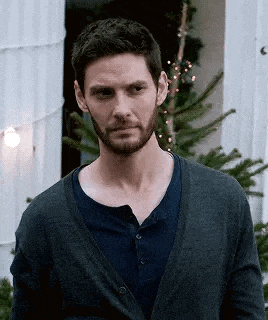 a man with a beard is standing in front of a christmas tree wearing a cardigan and a blue shirt .