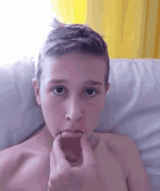a young boy without a shirt is laying on a bed with his mouth open and a hand reaching into his mouth .