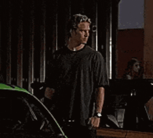 a man in a black shirt is standing next to a green car in a garage .