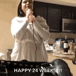 a pregnant woman is singing into a microphone in a kitchen while holding a bottle of wine .