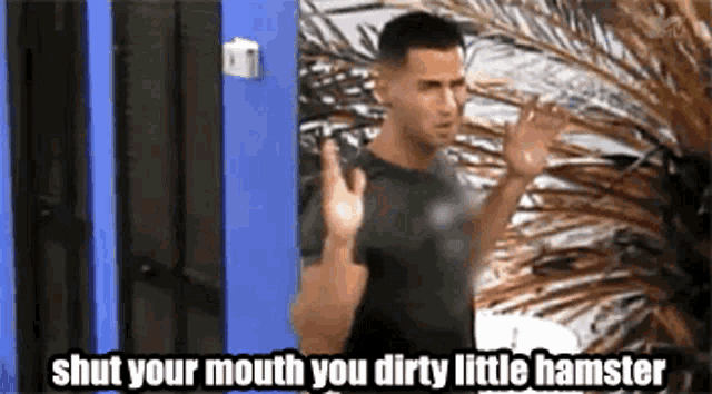 a man in a black shirt is standing in front of a blue door and says `` shut your mouth you dirty little hamster '' .
