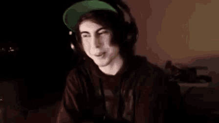 a young man wearing headphones and a green hat is smiling while sitting in front of a computer .